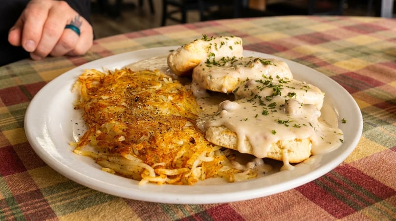A plate of golden hash browns and biscuits with creamy gravy, garnished with parsley, on a plaid tablecloth.