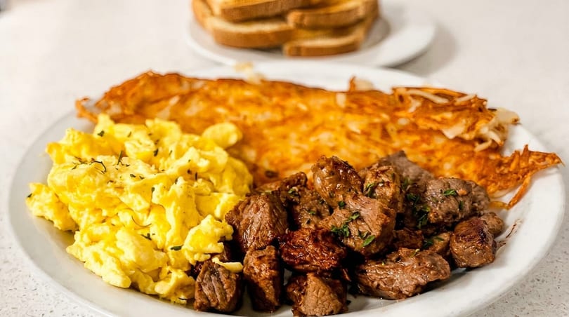 A breakfast plate with scrambled eggs, hash browns, and steak bites, with toast in the background.