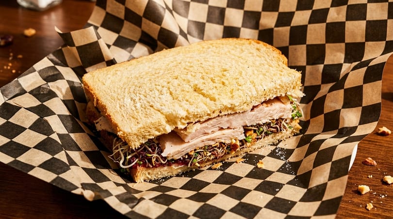 Turkey sandwich with sprouts and cranberry sauce on white bread, wrapped in checkered paper.