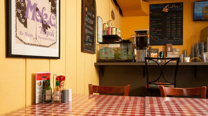 A cozy cafe interior with red and white checkered tablecloths, coffee bar, and menu boards.